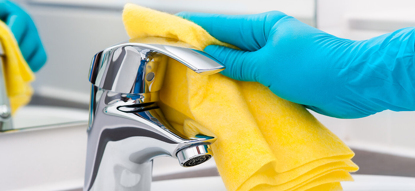 Hygienische Reinigung eines Wasserhahns mit blauem Handschuh und gelbem Wischlappen